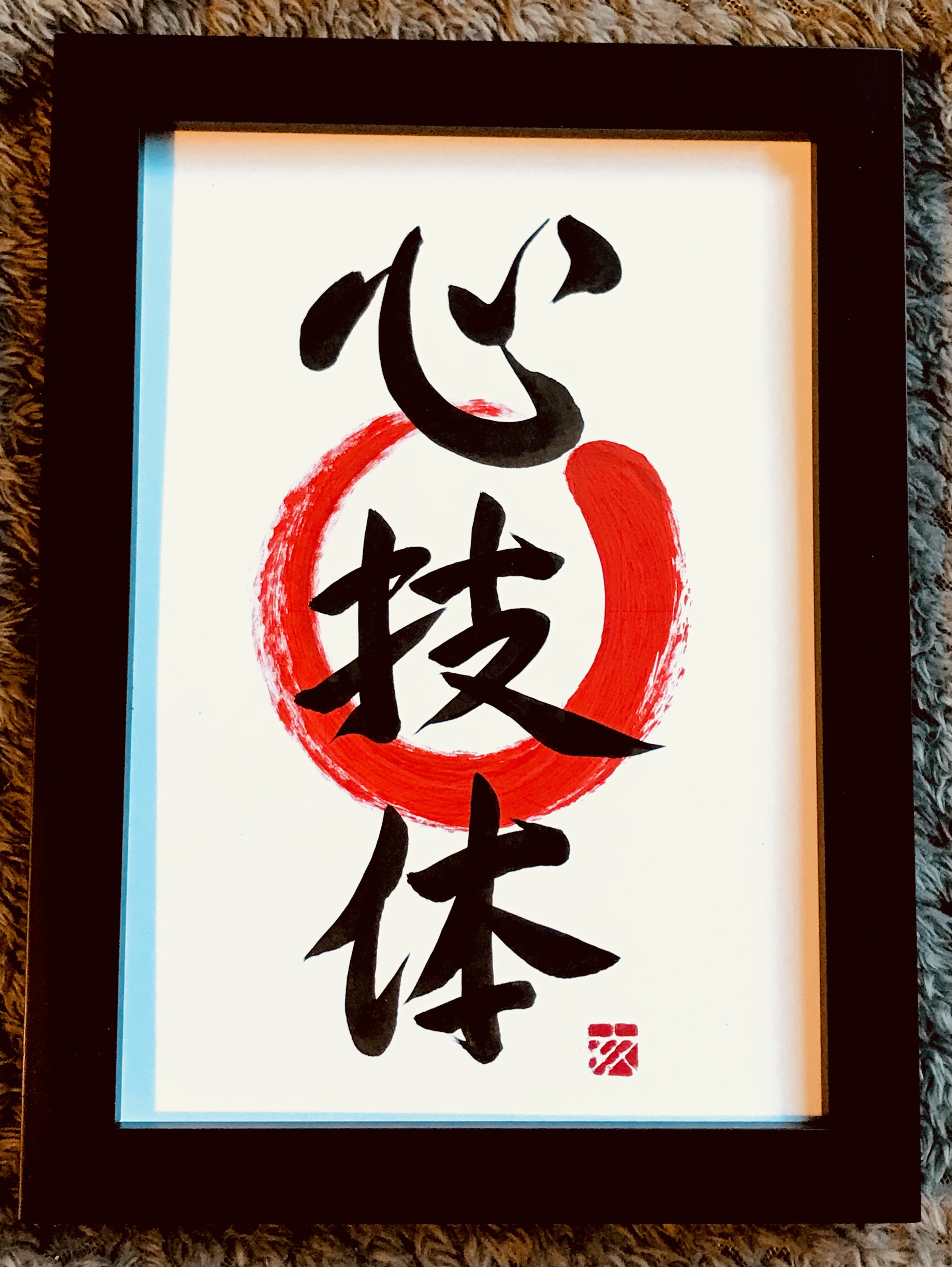 A Japanese calligraphy artwork of 3 characters, black ink on white paper. It means "heart, mind, body". A red enso (circle symbolising enlightenment) is behind the characters.