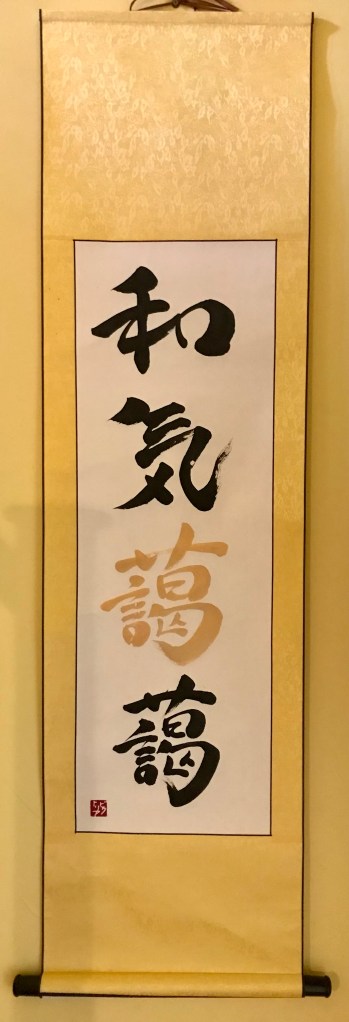 A Japanese calligraphy artwork of 4 characters written vertically on a traditional hanging scroll. Characters 1, 2 and 4 are in black ink. Character 3 is in gold ink. The 4-character phrase means "a happy home".