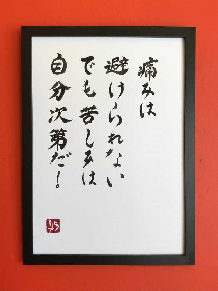 An A3-size Japanese calligraphy artwork. There are 4 vertical lines of black text on white paper. The work is framed and hanging on a wall.