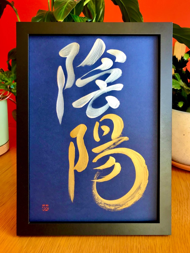 A Japanese calligraphy artwork of 2 characters. The top is in silver ink, the bottom is in gold ink. The paper is dark blue. It means "yin-yang".