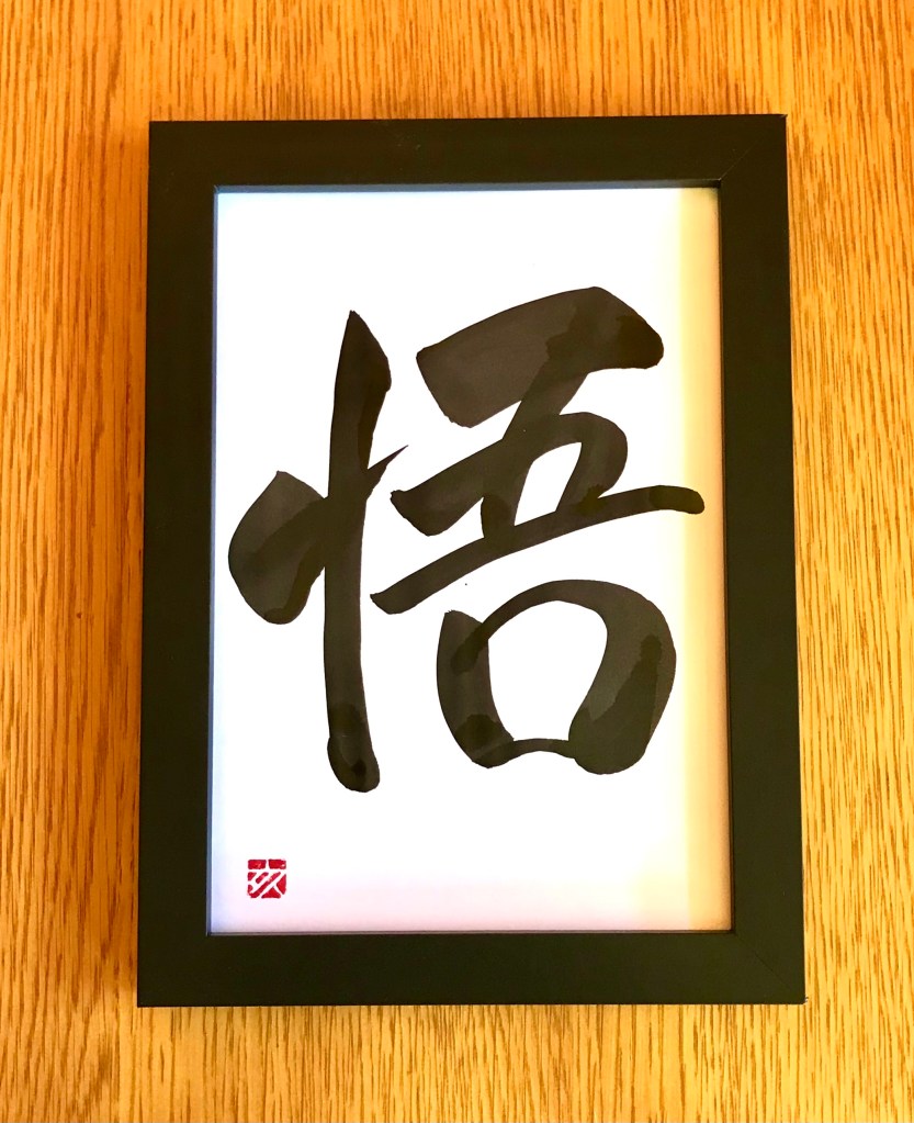 A Japanese calligraphy work of 1 character, black ink on white paper, in a black frame.