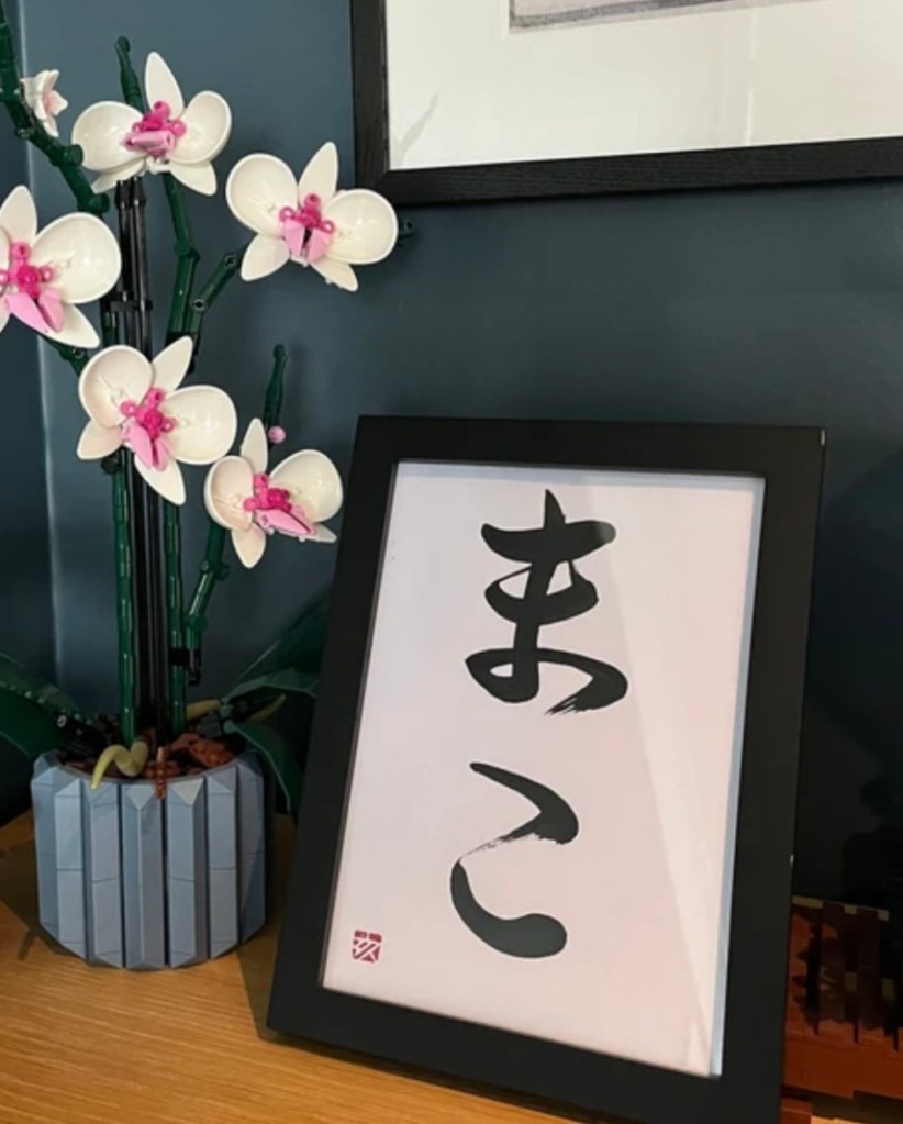 An A5-size framed 2-character Japanese calligraphy artwork, black ink on a white background. The artwork is displayed on a table next to an artificial plant.
