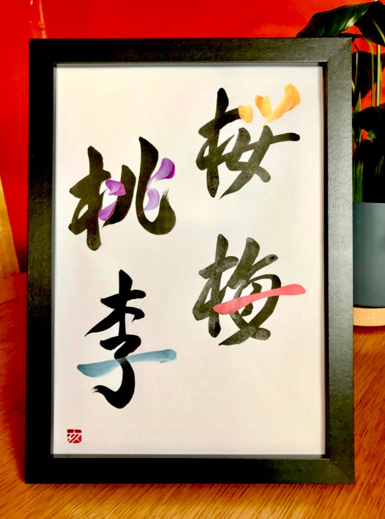 A Japanese calligraphy work of 4 characters, white paper in a black frame. The ink is mainly black, with some part of the characters written in coloured metallic inks.