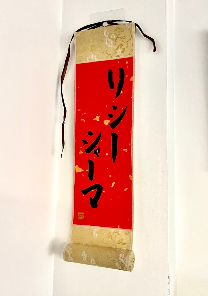 A small Japanese calligraphy artwork of black characters on a small hanging scroll. 