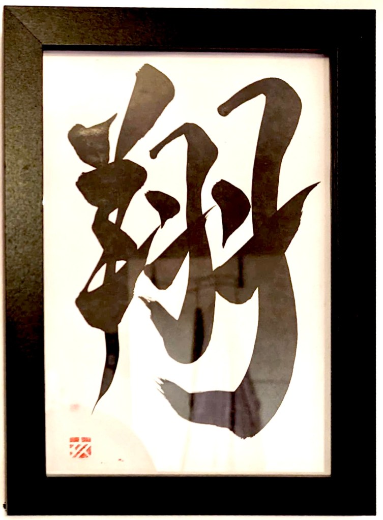 A Japanese calligraphy work of 1 character, black ink on white paper, in a black frame.