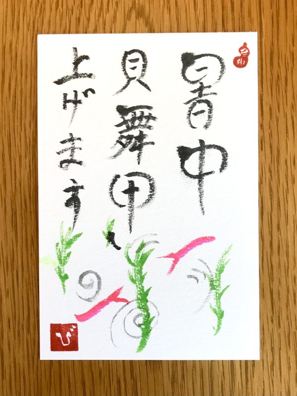 A postcard with Japanese characters for "summer greetings" written on it, with a simple image of some goldfish and reeds in red and green ink.