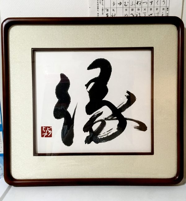 A framed Japanese calligraphy work of the character for "destiny" in black ink on a white page.