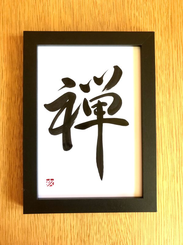 A 1-character Japanese calligraphy work, black ink on white card, in a black frame.