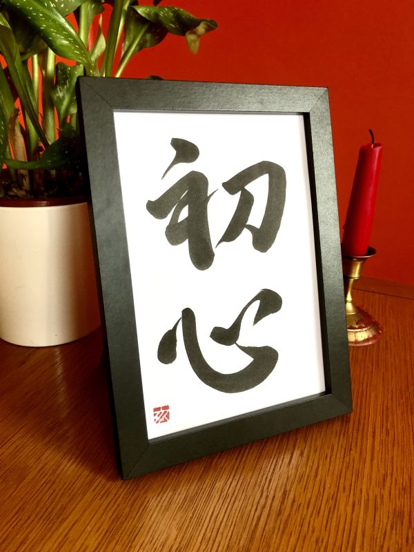A 2-character Japanese calligraphy work, black ink on white card, framed on a table. A plant and candle are in the background.