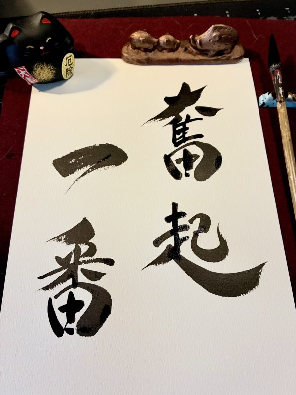 A Japanese calligraphy artwork of 4 characters, black ink on white text. The artwork is on a desk with a brush next to it and gold paperweight at the top.