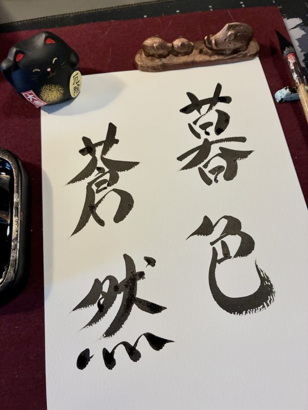 A Japanese calligraphy work of 4 kanji characters. The characters are in black ink on white card. There is a brush to the right side and an ink stone to the left side. At the top is a paperweight and a small cat ornament.