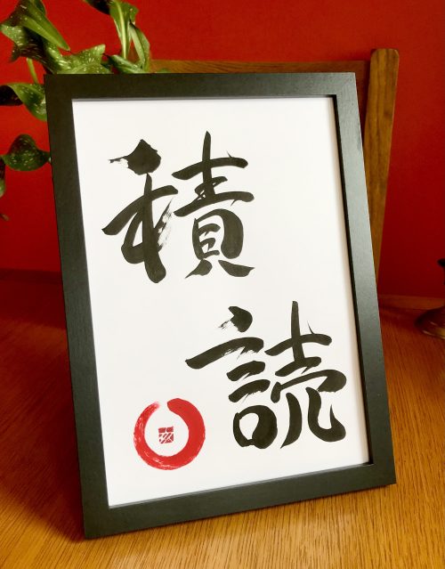 A framed Japanese calligraphy work of two characters, black ink on white card. In the bottom left corner is a small red "enso" (an incomplete circle).