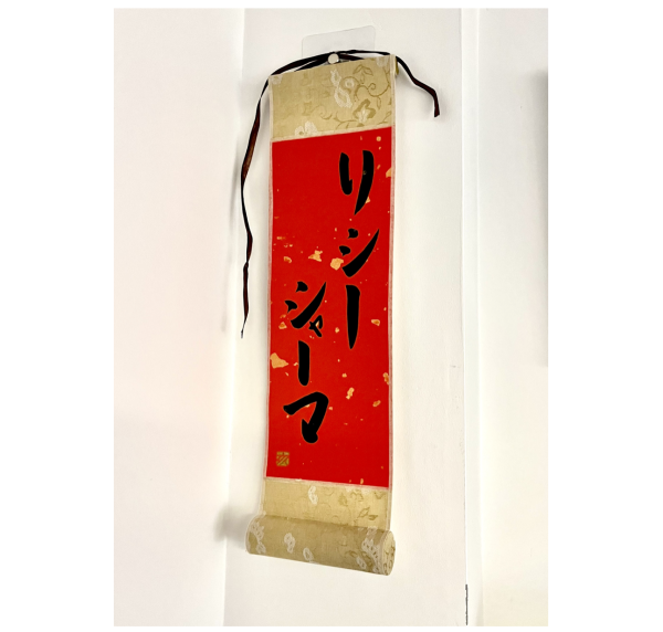 A Japanese calligraphy work in black ink on a small red hanging scroll. The characters say the client's name.