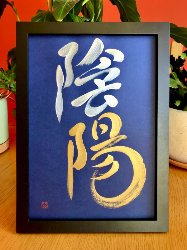 A Japanese calligraphy work  in silver and gold ink, on a dark blue background. The characters say "yin-yang". The work is in a black frame.